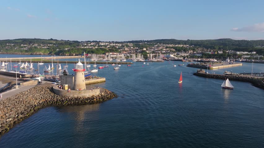 Sail craft and pleasure boats navigate the channel beside Howth Pier lighthouse with the marina and town in bright midday light.