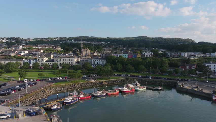 Fishing boats line a small basin beside a green seafront park and hillside houses on a bright summer day, viewed from a rising drone.
