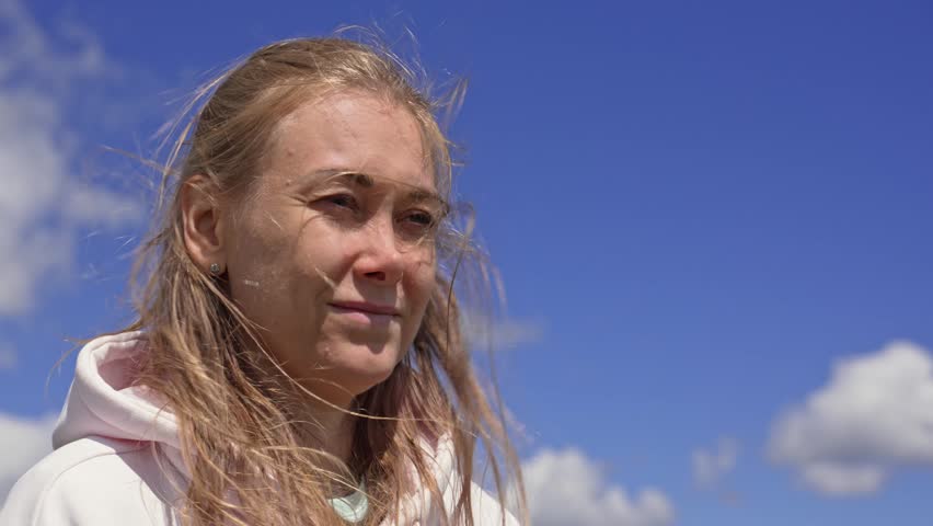 Beautiful blonde woman enjoying the sun and wind as her hair blows across her face against a bright blue sky with scattered clouds, symbolizing freedom, tranquility, and natural beauty