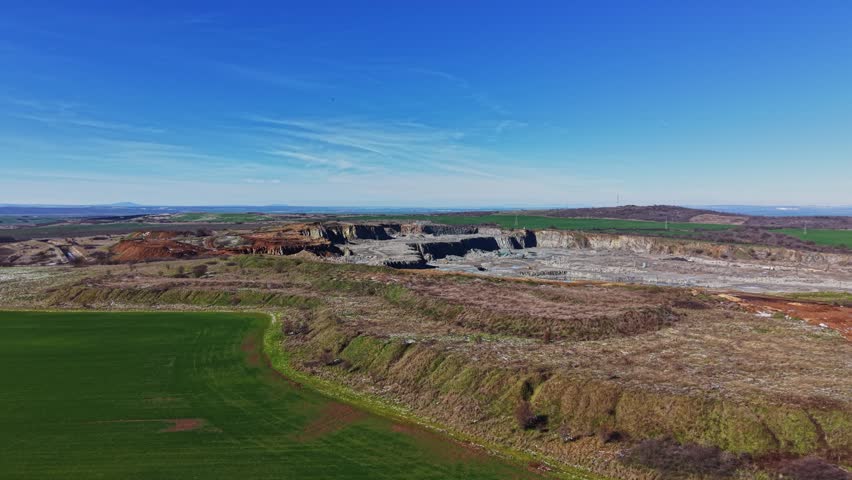 A large mining site shows excavation and land alteration with a water body present. The sky is clear and blue. Surrounding fields are visible in the background near the site.
