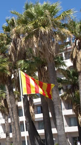 A red and yellow striped flag flutters before palm trees and a sunny hotel.