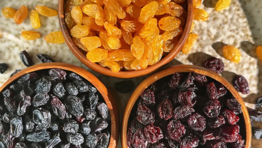 Overhead rotating display of three raisin varieties in wooden bowls showing dark red black and golden dried fruit