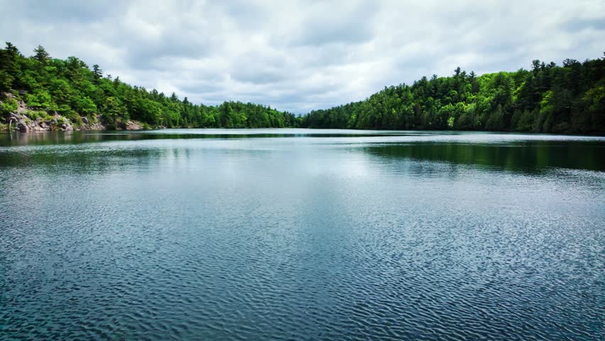 Wide panoramic landscape of Pink Lake (Lac Pink) in Gatineau Park near Ottawa, Quebec, Canada. Calm water surrounded by dense forest beneath dramatic clouds.