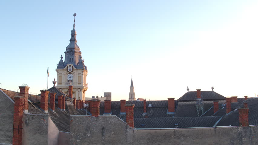 Aerial shot rising above old tiled rooftops to reveal the historic City Hall clock tower and city skyline of Cluj-Napoca under a clear blue morning sky