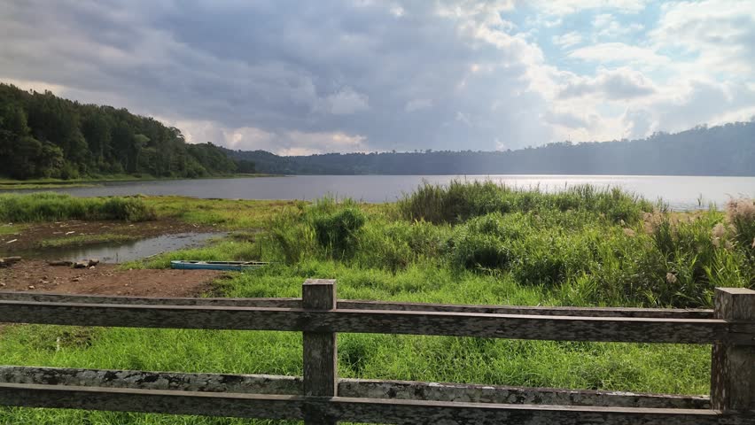 Scenic Lake View with Wooden Fence and Green Meadows Under Cloudy Sky