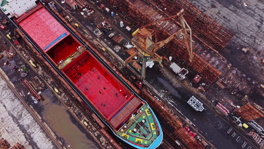 A large vessel is positioned at a shipyard. Workers are busy loading and unloading cargo as heavy machinery operates nearby. The area is full of materials and equipment.