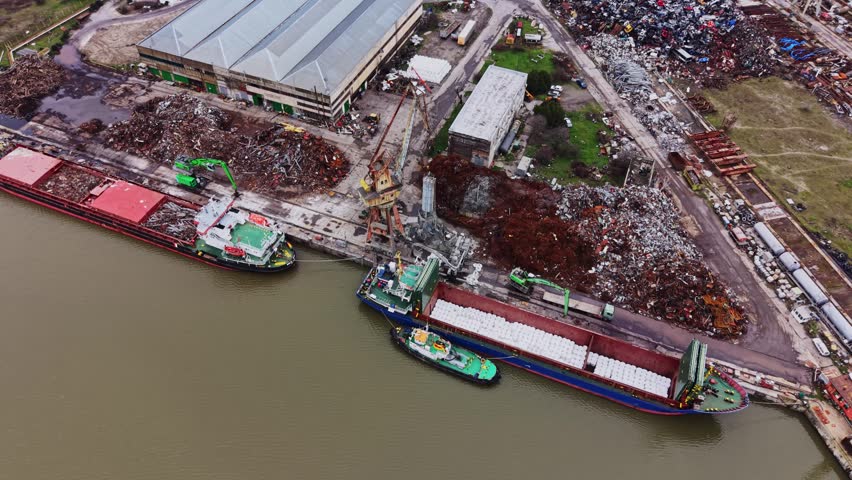 Cargo vessels are docked at a shipyard. The cranes are loading items onto the ships while nearby, piles of scrap metal are stored. Trucks are waiting for transport.
