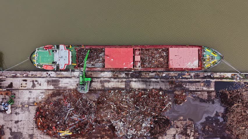 A cargo vessel is docked at a shipyard beside a river. Workers are unloading waste material from the vessel onto the dock. The surrounding area has debris and equipment.