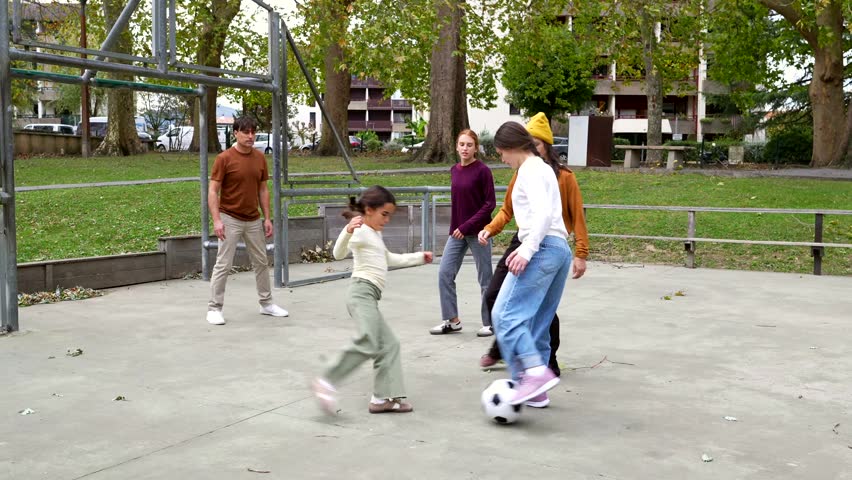 Cheerful group of friends enjoying a casual soccer game on a concrete court in a city park. Young women and children are actively playing together, kicking the ball and running outdoors