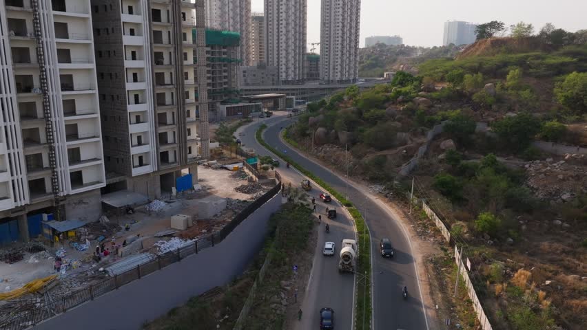 Vehicles moving on S shape curved road with under construction tower building and rocky terrain at puppalguda, manikonda, hyderabad, telangana, india. day time, stable shot, drone shot, 4k.