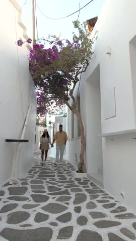 A couple strolls through the beautiful, narrow streets of Naousa, Paros, Greece, where blooming flowers add color to whitewashed buildings. Enjoy the charm of the Cyclades 