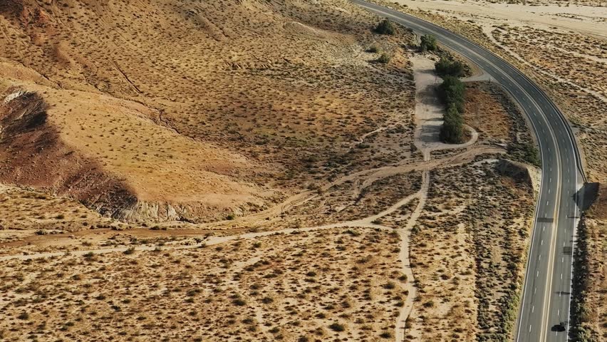 A winding road cutting through dry land in the USA. The area is mostly barren with a few patches of greenery near the road. It highlights the landscape and terrain.