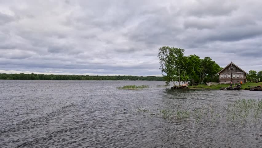 A lone wooden cabin stands by a vast lake under a dramatic cloudy sky. Reeds and grasses line the calm rippling shore.