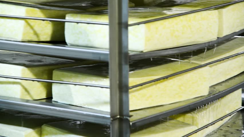The washing process during cheese production at a cheese factory. A metal shelf with large rectangular blocks of pale yellow cheese. Cheese is at different stages of processing and production.
