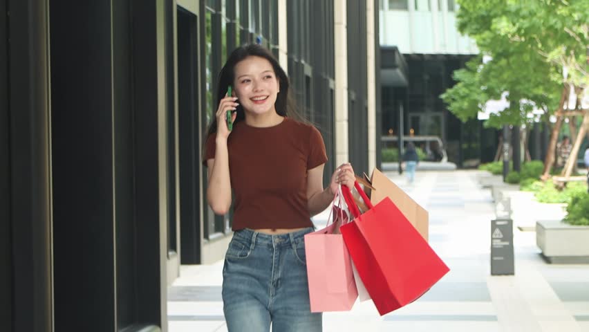 Cheerful young girl enjoys a shopping in city. Beautiful Asian woman with shopping bags using mobile phone during walking in downtown. Happy girl talks on smartphone while strolling through the mall.
