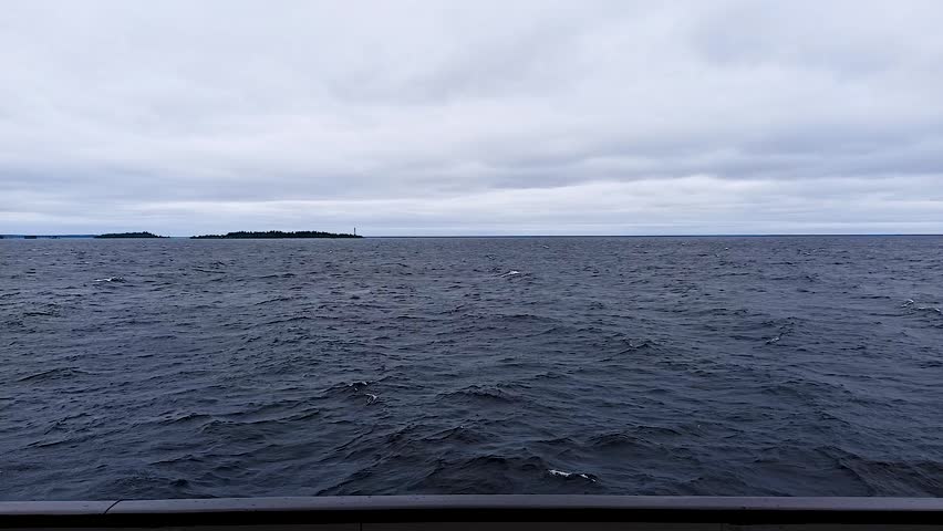 A huge stormy sea stretches under an overcast sky, with a distant tree-covered island and a lighthouse on the horizon. Dark undulating water and the edge of the ship
