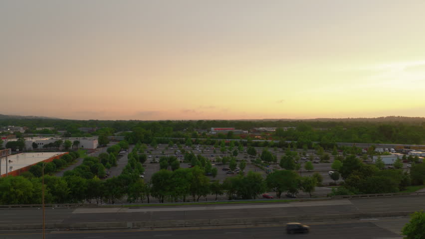 Wide aerial shot of cars driving on a highway next to a tree-lined commercial parking lot in a Birmingham, Alabama suburb during a warm golden hour sunset.