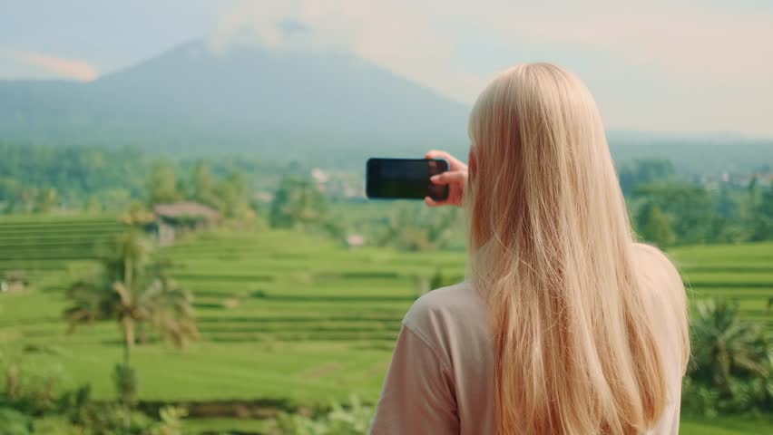 A blonde woman photographed from behind taking a picture of a volcano and rice terraces on a bright sunny day. She stands facing the scenic landscape outdoors.