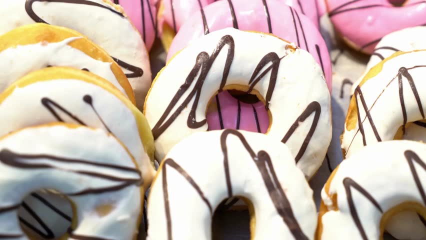 Several delicious donuts fill the screen. Donuts are topped with white glaze and drizzled with dark chocolate. Some donuts have pink frosting with chocolate stripes.
