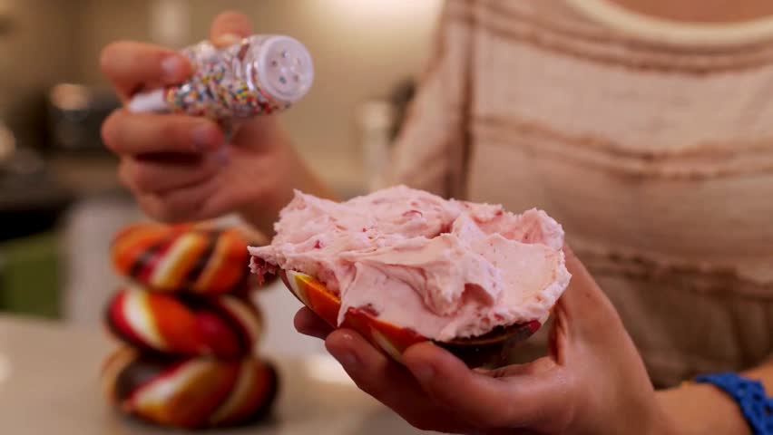 Young Woman decorates donut with light pink frosting and colorful sprinkles. Delicious snack being prepared. Stack of colorful donuts in background.