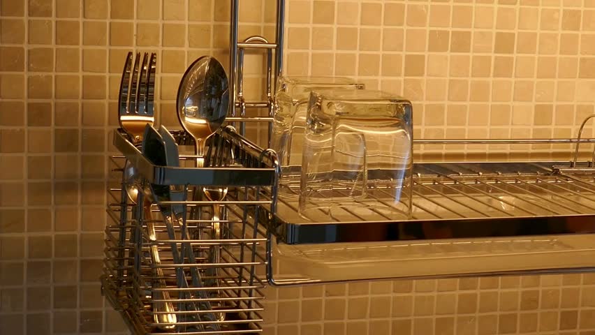 Hand places clean glassware and white plates onto drying rack in contemporary kitchen. Bright, warm lighting highlights neat arrangement of dishes.