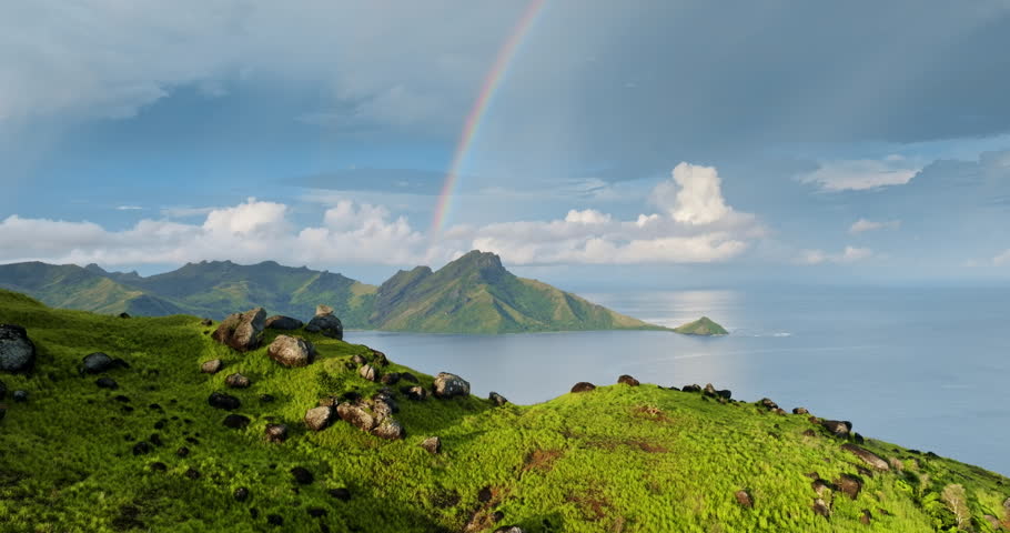 Amazing Fiji paradise landscape. Lush green hills with scattered rocks overlooking a serene ocean bay with tropical islands and mountains under a bright rainbow arching the blue sky with clouds