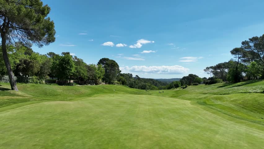 Expansive view of well-maintained golf course featuring vibrant green fairways, surrounded by trees and distant hills under a bright blue sky.