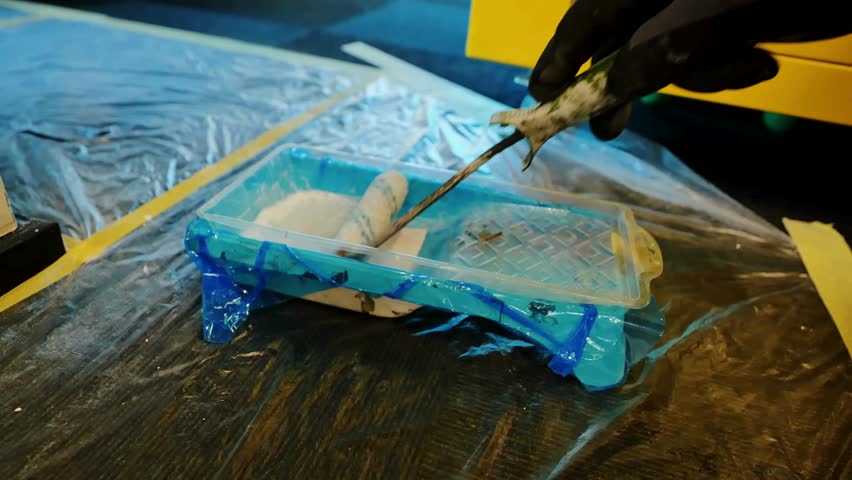 Roller brush moves across paint tray filled with white paint on clear plastic covering floor in a workspace. Bright yellow cabinet stands in background.