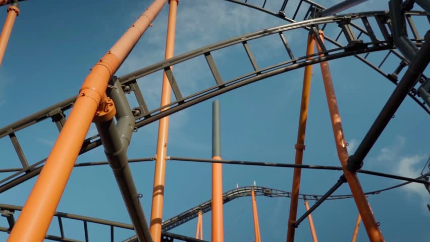 A close view of orange roller coaster tracks intertwining against a clear blue sky. Discover the excitement of amusement parks and the thrill of roller coasters in this dynamic setting.
