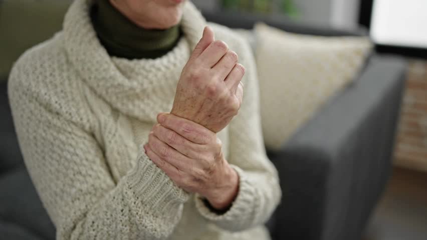 Woman in modern living room grips her wrist with discomfort. She wears a warm sweater. Background includes a couch and decorative pillows. Lighting is soft and bright.