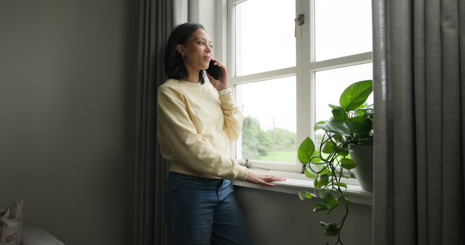 African American woman leaning on sill, receiving call, talking on phone by multi-pane window. Portrait, candid, home, plant, curtains, smartwatch, casual