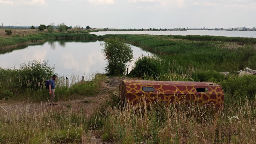 Back view of a man with a backpack walking down a dirt path towards an abandoned camouflage military wagon near a lake