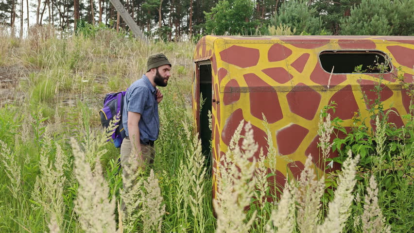 Man with a backpack standing at the open entrance of an abandoned camouflage military wagon and looking inside