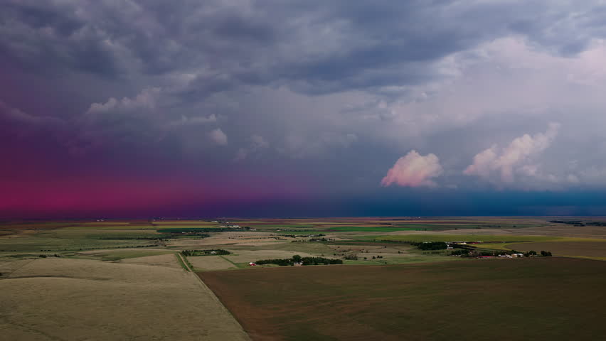 Lightning illuminates towering storm clouds above patchwork farmland as warm sunset colors fill the sky, creating a dramatic aerial landscape with powerful weather and striking natural contrast.