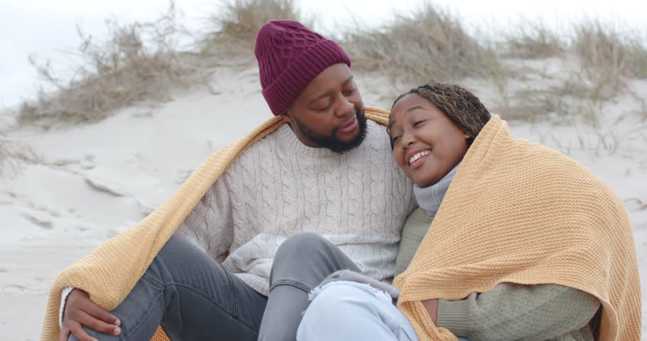 African American couple sitting on dune, man draping mustard blanket, seeking warmth and closeness. Romance, intimacy, cozy, eyecontact, outdoor, beach, sand