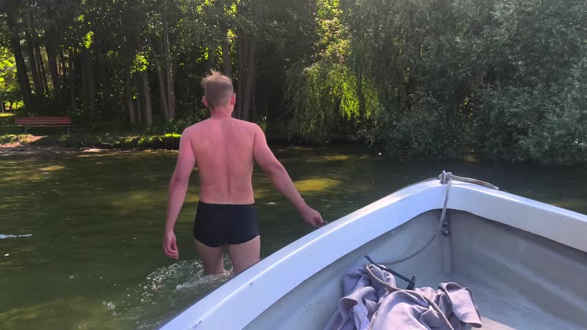 Man in swimming trunks walking in shallow water and pulling a boat with a rope to the shore