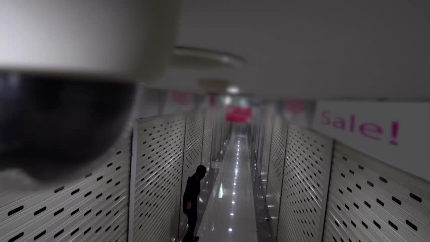 Surveillance camera captures an empty shopping mall corridor with a single figure walking quietly. Lights reflect on polished floors creating a focused atmosphere.