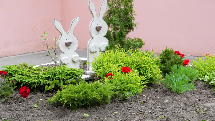 Two handmade white wooden rabbit figurines stand inside a cultivated garden bed surrounded by green shrubs and blooming red flowers against a pink wall.