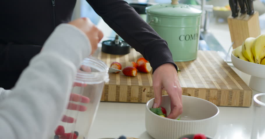 Diverse women friends trimming strawberry stems on board, adding yogurt to blender cup for smoothie. Berries, fruits, kitchen, knife, leaves, bananas, blueberries
