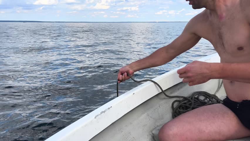 Close-up of a man pulling a rope to lift a folding grapple anchor out of the lake water into a boat