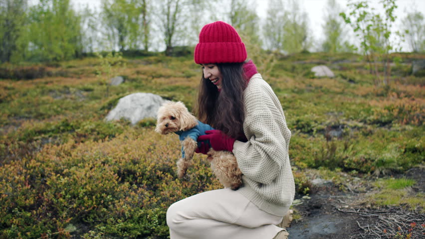 A smiling young woman in a knit hat and beige sweater kneels on a rocky green hillside while gently holds and throws up a small fluffy terrier dog. A moment of love and affection for a pet
