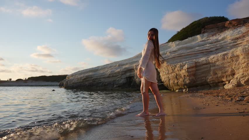 Happy young woman walking barefoot in the sea along a sandy beach. She is joyfully raising her arms, celebrating freedom and nature