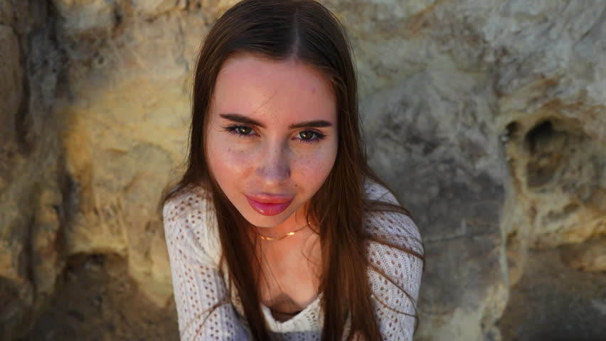 High angle portrait of a beautiful young woman with long brown hair and freckles playfully posing and smiling at a rocky beach during sunset