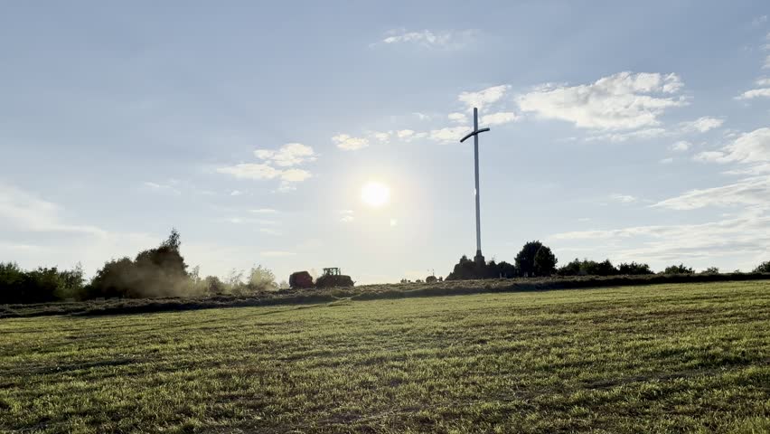 A scenic wide shot of a red tractor baling hay on a green agricultural field under a beautiful partly cloudy sky. In the foreground, several round hay bales lie scattered across the mown grass, while a tall, modern Christian cross monument stands on a hill nearby, blending farming life with spiritual symbolism.