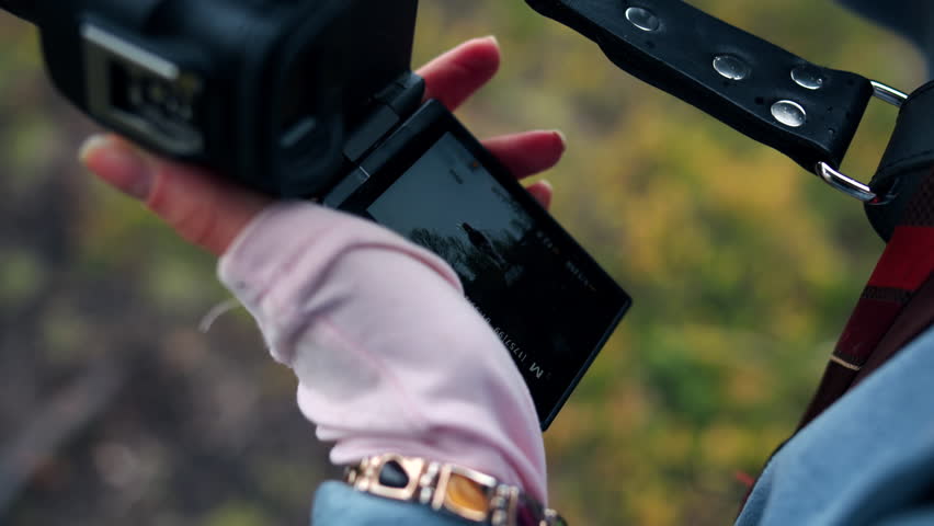 Close-up of a left female hand in a pale pink glove holding a mirrorless camera attached to a leather belt wit flip-out LCD screen tilted toward the viewer. The photographer is taking series of photos