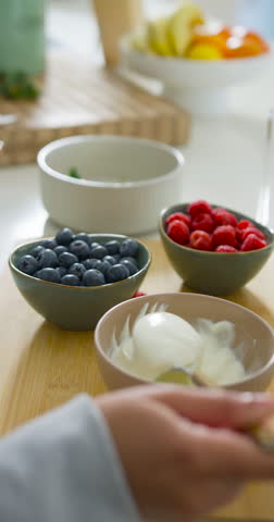 Vertical video: Diverse women spotting bowls, scooping yogurt into blender cup, prepping smoothie. Kitchen, counter, fruits, berries, dairy, teamwork, vertical video