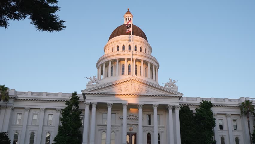 The California State Capitol stands tall as the sun sets behind it in Sacramento, capturing warm lights against the evening sky.
