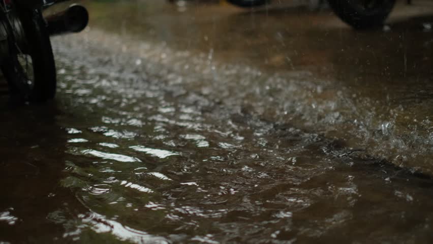 Raindrops splash on wet pavement near parked motorcycles. Water ripples across dark, reflective surface. Rain creates dynamic patterns on asphalt. Moody atmosphere captures rainy day intensity