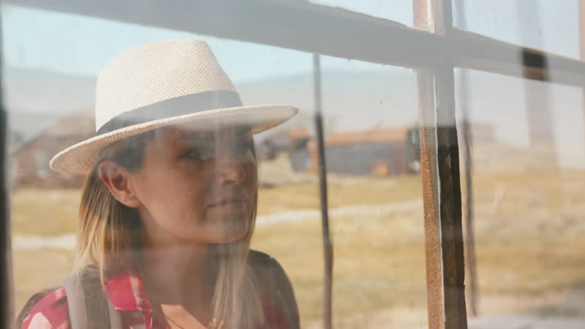Girl walks through old ghost town in America peering through dusty window of abandoned building. Woman in straw hat and plaid shirt touches weathered glass pane in rural desert setting.