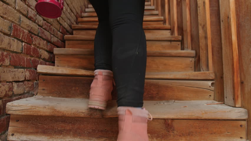 Woman in black leggings and pink sneakers climbs rustic wooden staircase in old brick building, carrying vintage red lantern. Low angle view captures worn timber steps and textured walls.